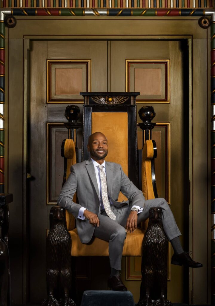 Photo of a black man in a suit sitting in a chair in front of large doors