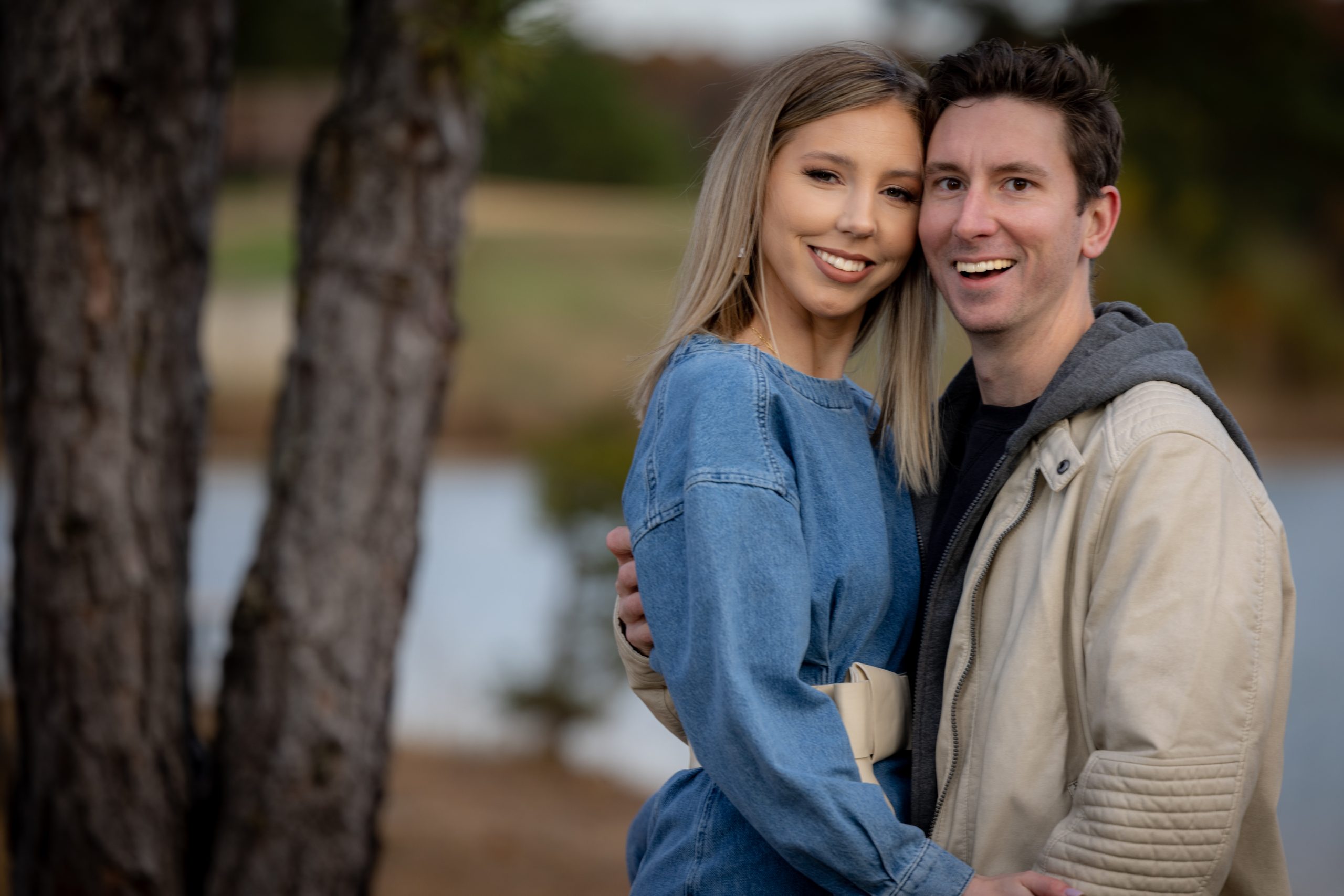 joyful engaged couple laughing by lake with fall foliage in the background