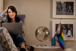 woman on computer with globe, family wall portraits, and pride flag