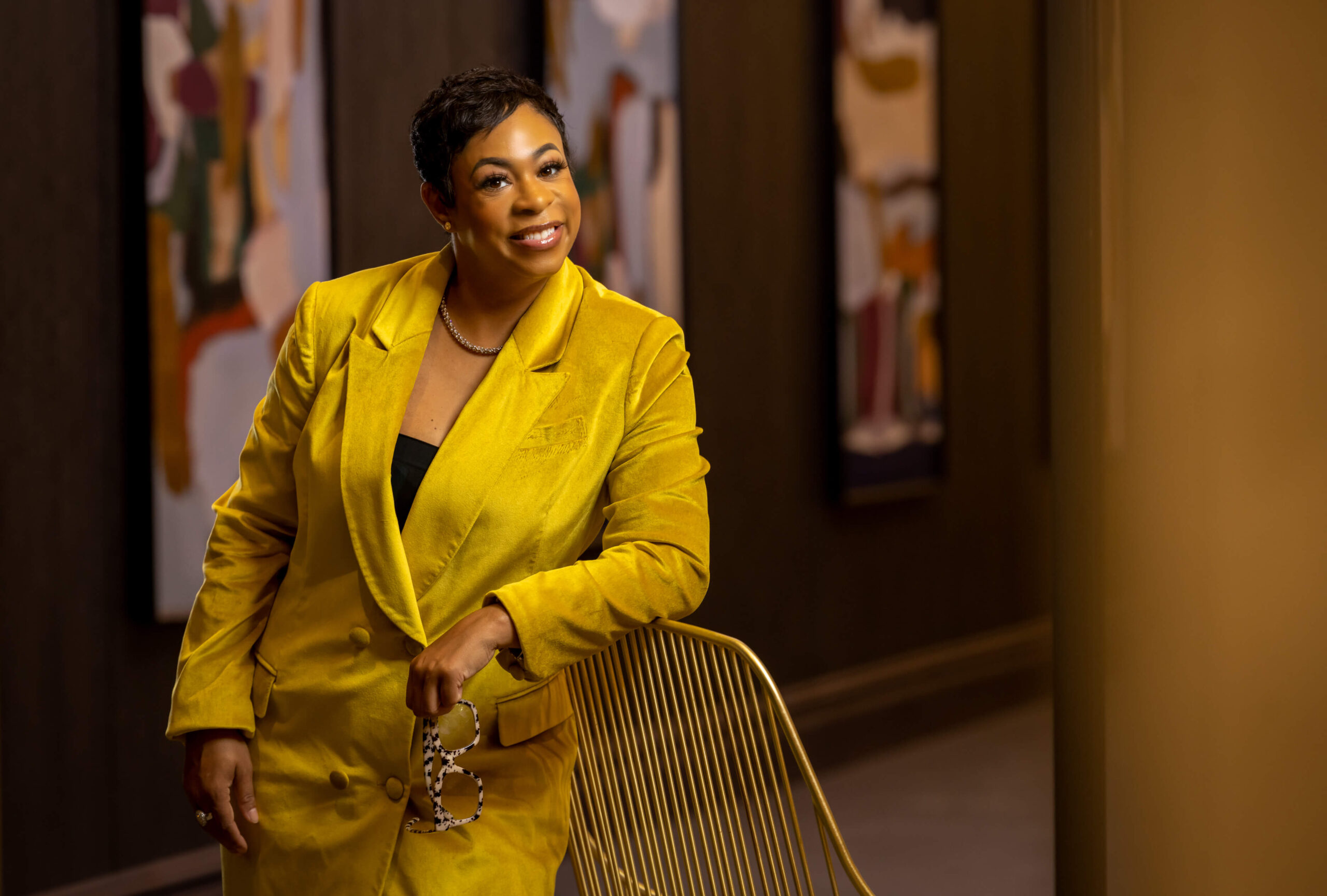 cool woman leaning on a chair while wearing a yellow suit and holding glasses