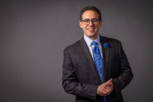 man in blue and grey suit clasping hands while smiling