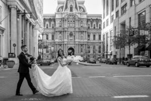 groom holding bride's wedding dress as they walk by broad street at city hall in philly