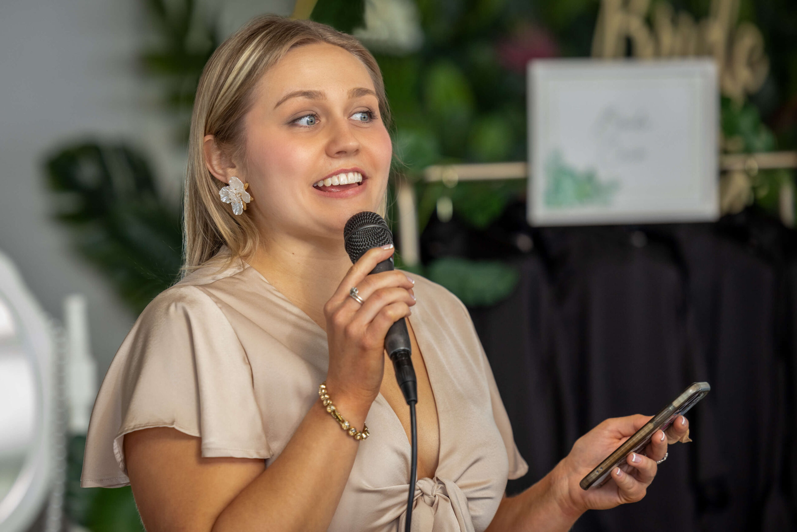 blonde woman holding a microphone and phone to give a speech at a bridal shower