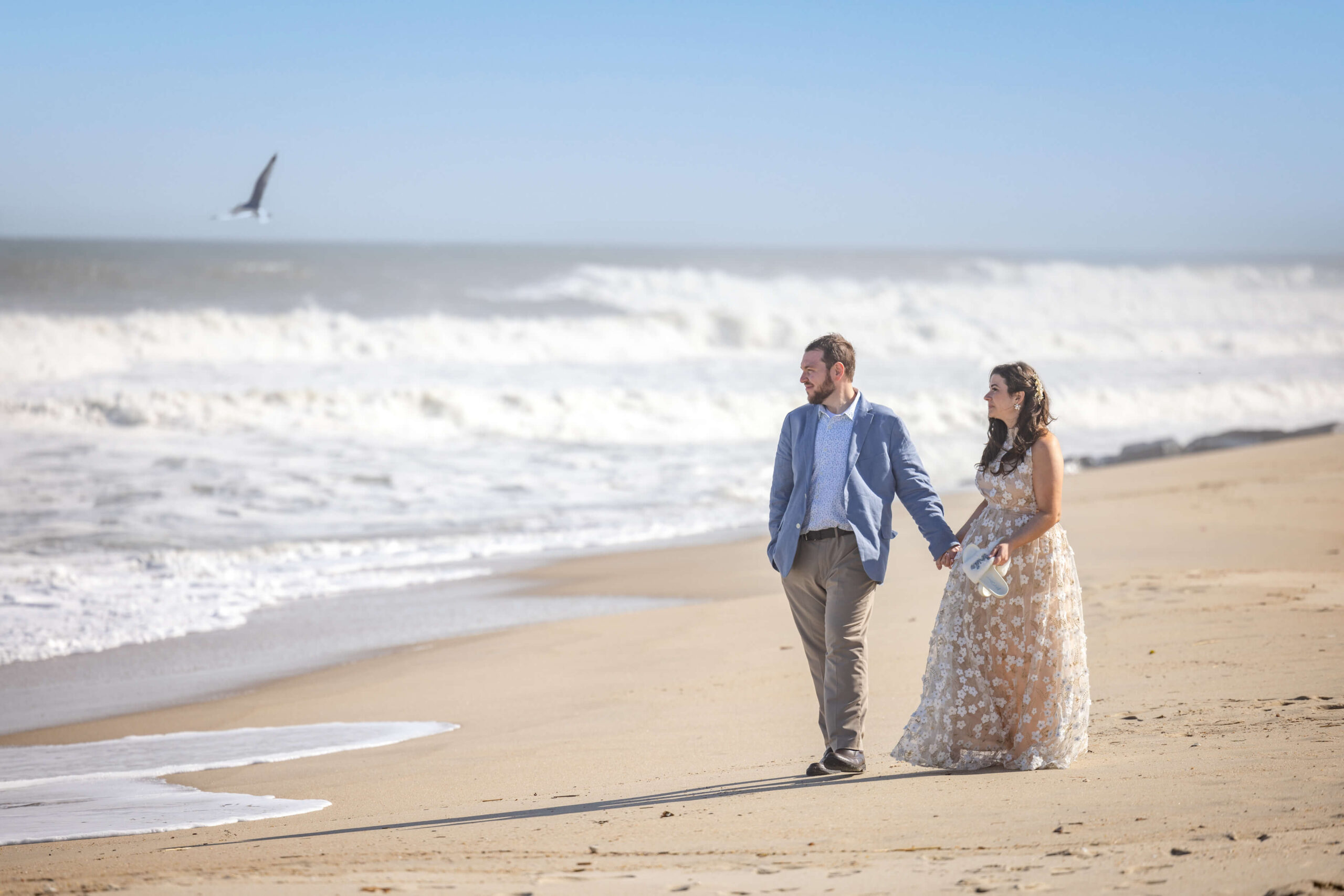 engaged couple hold hands and walk on the beach as a seagull passes overhead