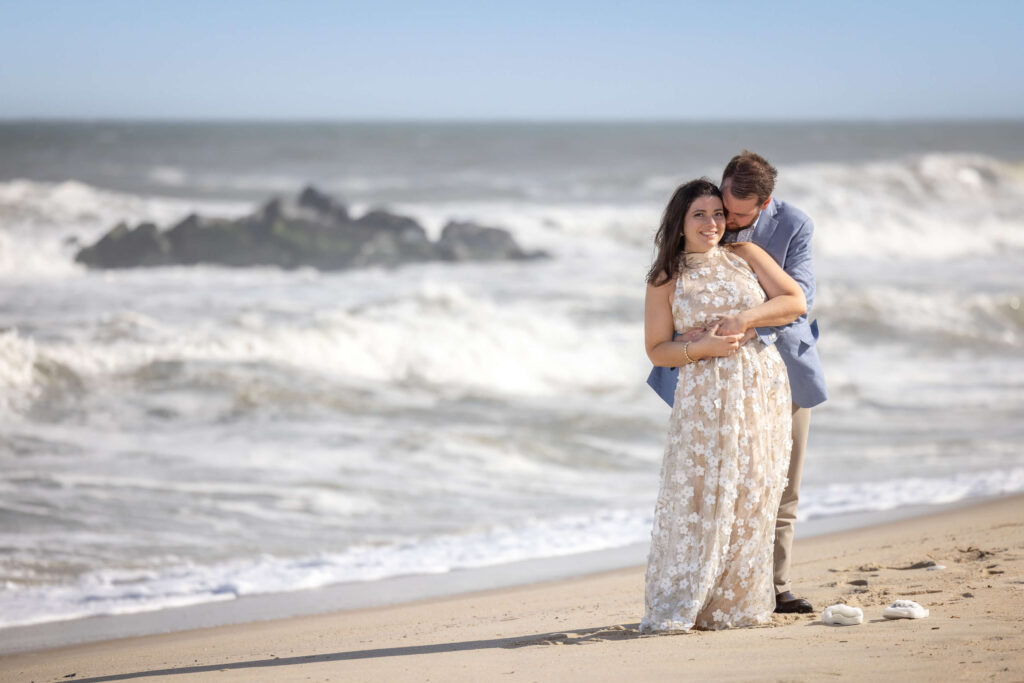 couple embracing on the beach as waves crash against the rocks on clear sunny day