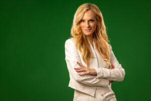 blonde woman in ivory white jumpsuit crossing arms against green background