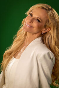 blonde woman wearing an ivory white outfit and smiling and tilting head against green background