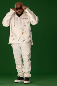 man in white hoodie, white ripped jeans and black shoes putting sunglasses on against green background