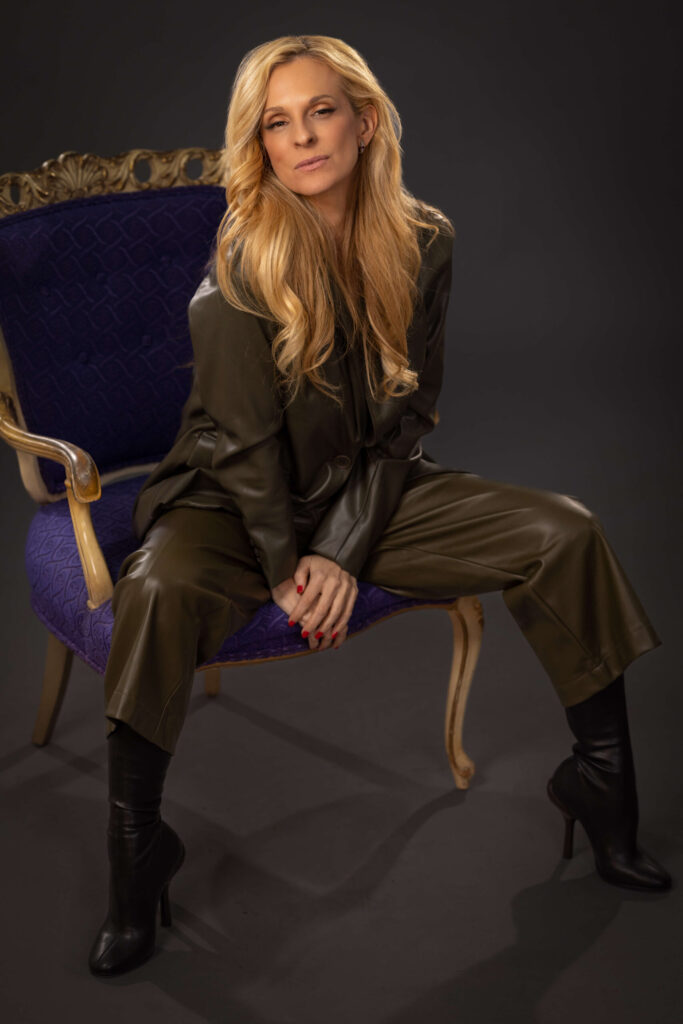 blonde woman in dark green outfit sitting in elegant purple chair against a grey background