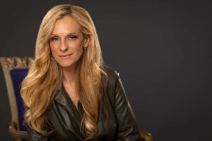 blonde hair women smirking in dark green top sitting in a purple chair against a grey background