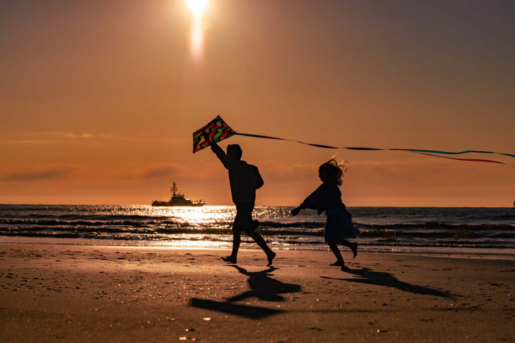 boy and girl playing on the beach during golden sunrise with a kite while fishing boat sails behind