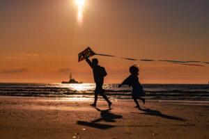 boy and girl playing on the beach during golden sunrise with a kite while fishing boat sails behind