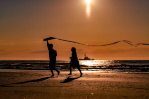 children running with a kite on the beach during golden sunrise with boat in background