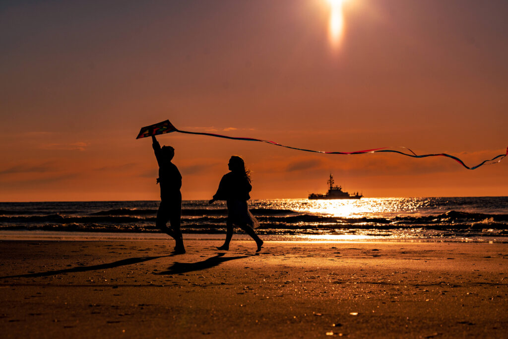 sunrise photo of children running on the beach with a kite, and boat in the background