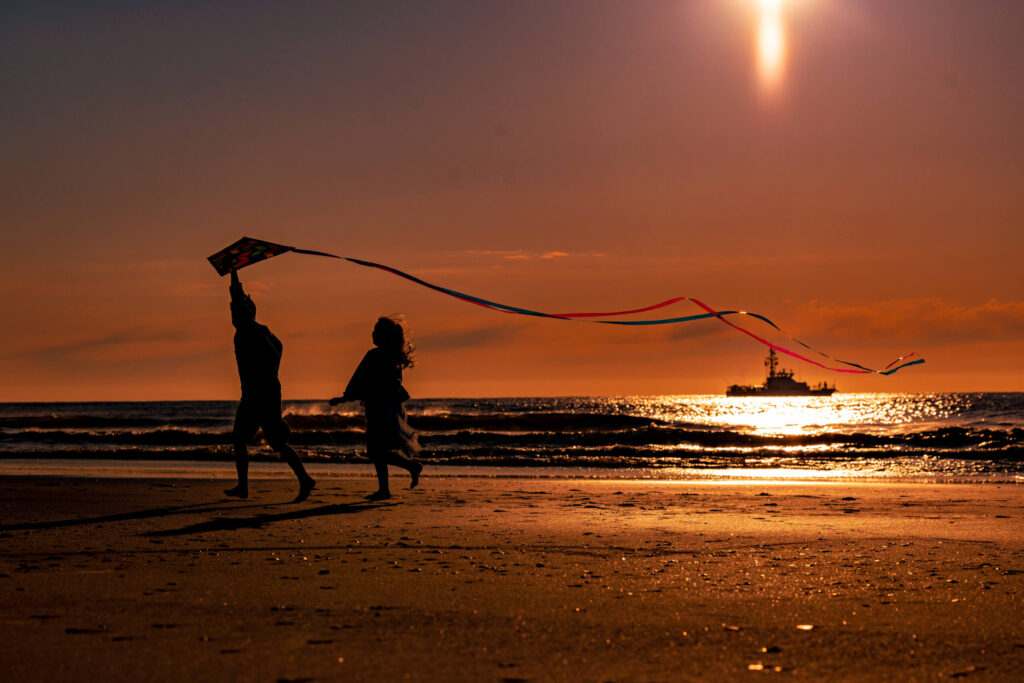 two young children running on beach with kite and fishing boat in ocean