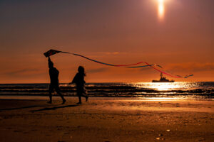 two young children running on beach with kite and fishing boat in ocean