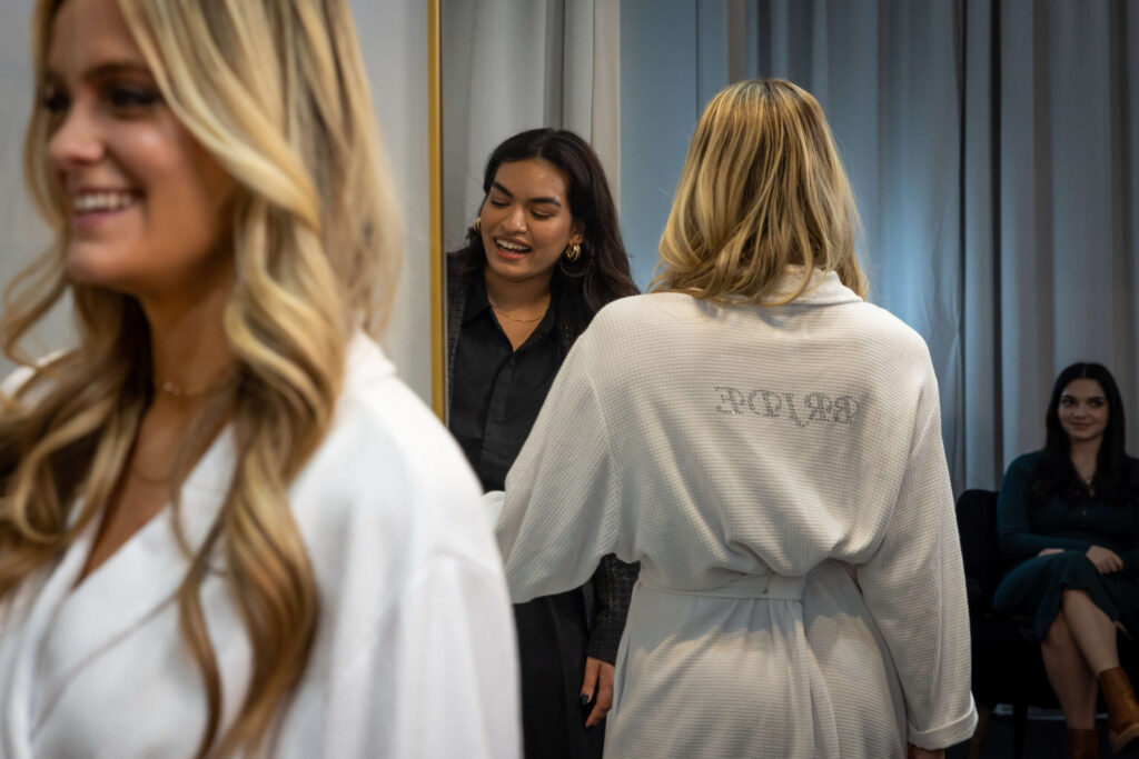stylist talking to a blonde woman in white robes with silver letters that read "bride" on the back of robe