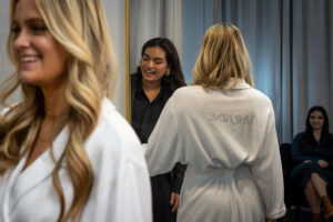 stylist talking to a blonde woman in white robes with silver letters that read "bride" on the back of robe