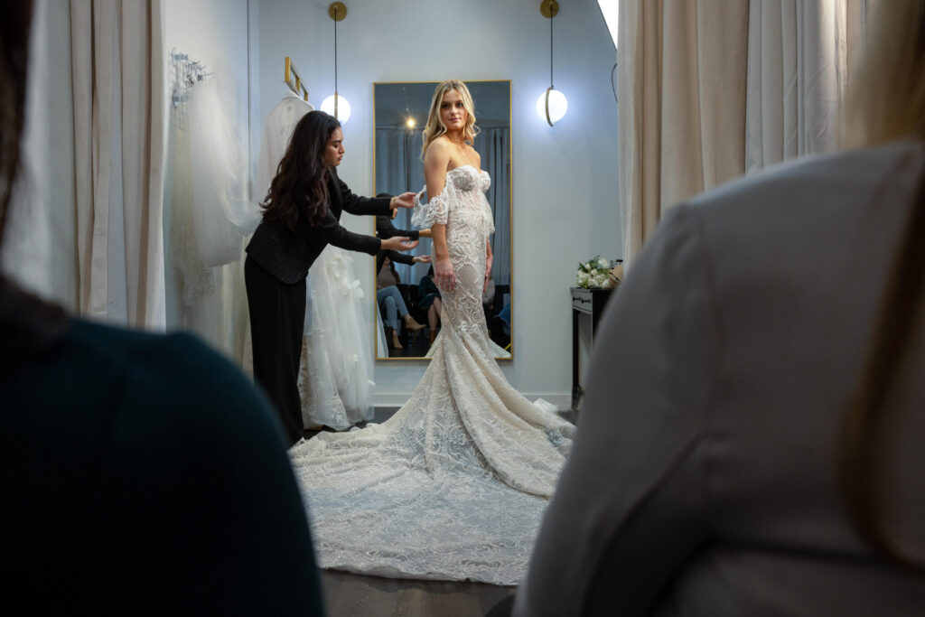 stylist touch-ups a customer's wedding dress as she poses for family and friends in changing room at bridal salon