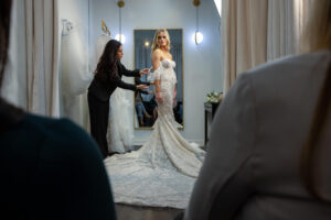 stylist touch-ups a customer's wedding dress as she poses for family and friends in changing room at bridal salon