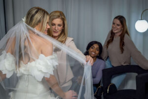 blonde woman holds a bride's wedding veil as friends smile from the side
