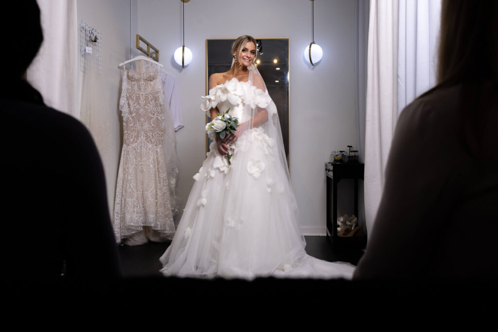 bride in changing room smiles for family and friends as she holds a white flower bouquet