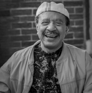sherman hemsley smiling while wearing a white hat and khaki jacket against a brick wall