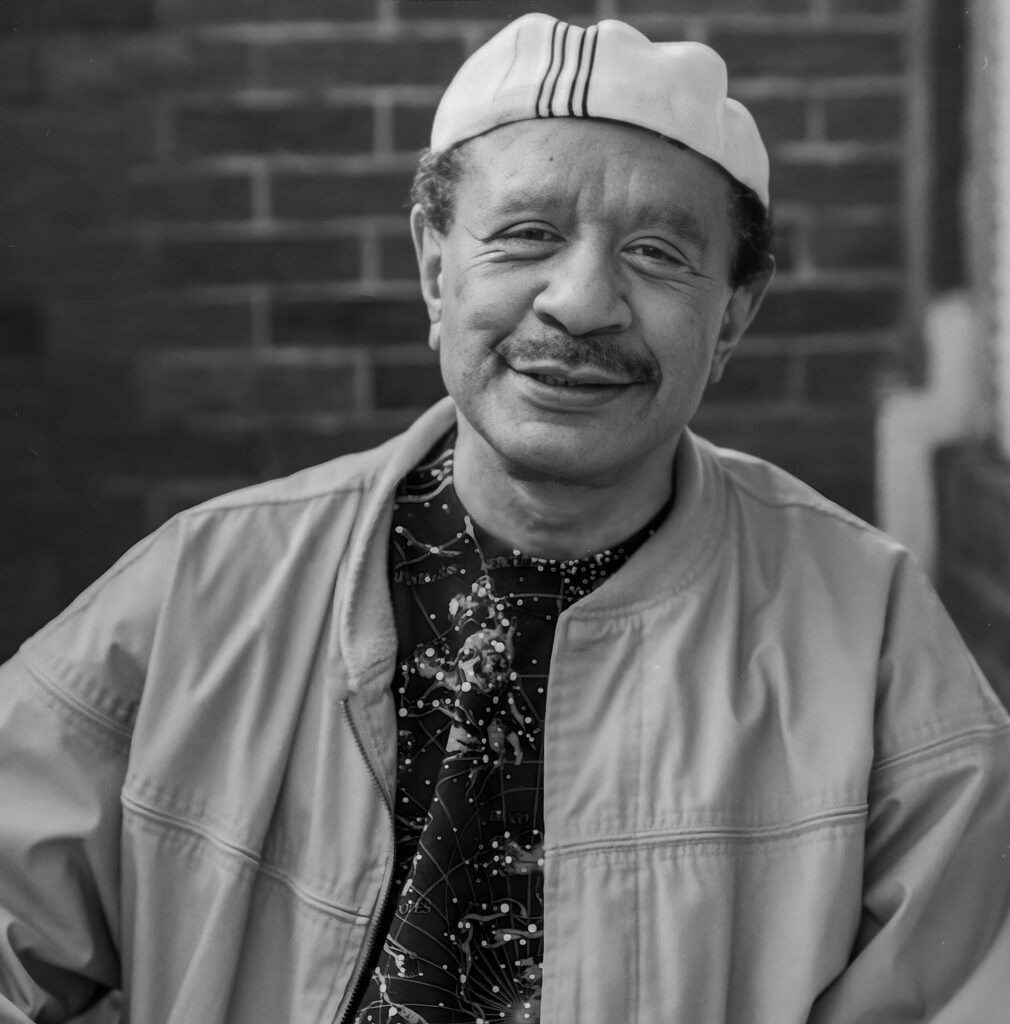 sherman hemsley posing against brick wall with backwards white hat and khaki jacket