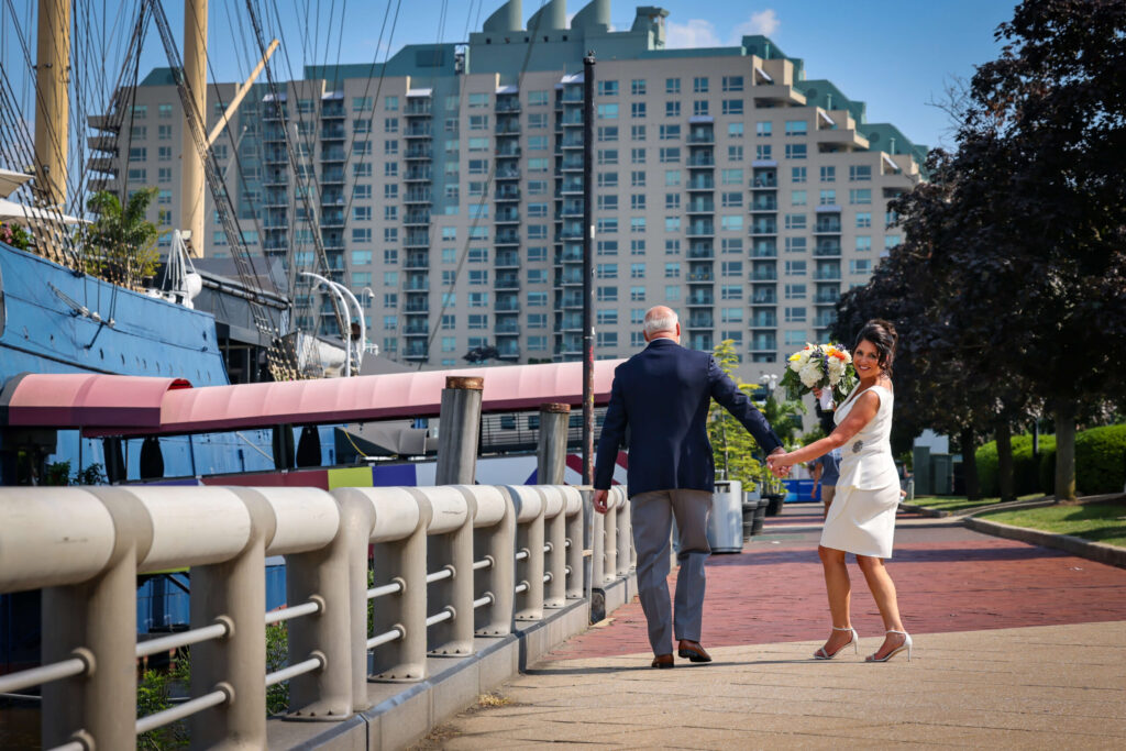 casual bride and groom walk by the moshulu in philadelphia on a sunny day. the bride looks back while holding a bouquet of flowers
