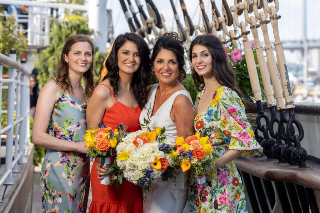 four women posing in colorful dresses while holding flower bouquets at the moshulu