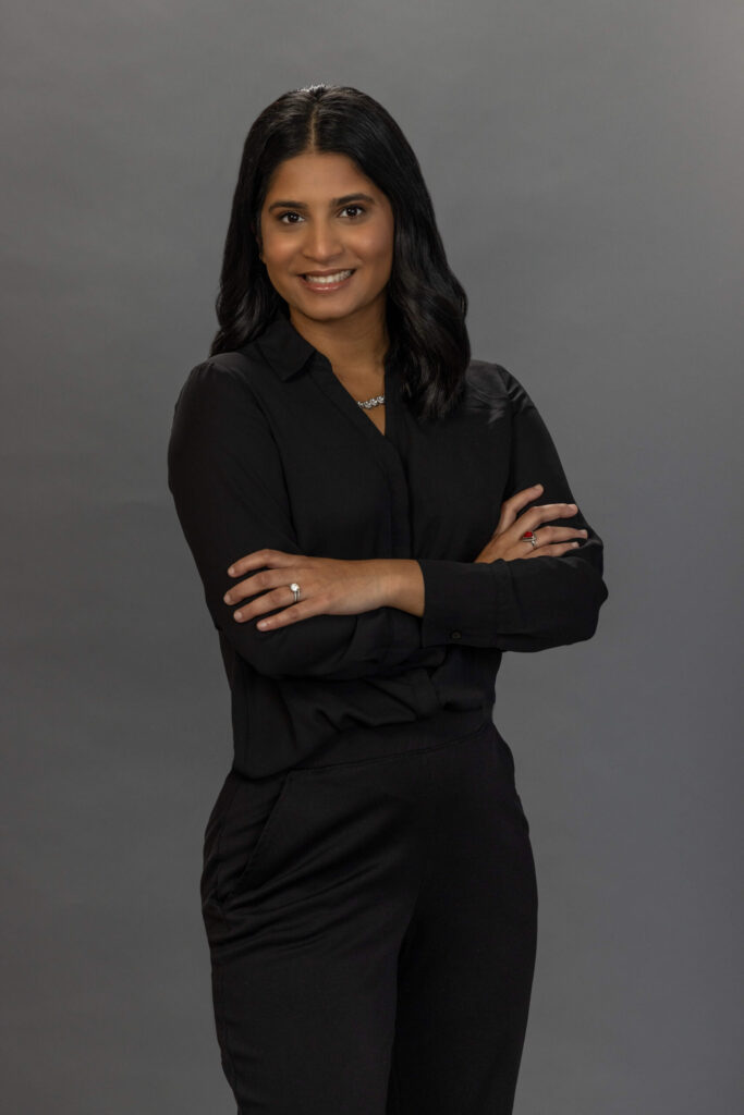 woman in all black outfit crosses her arms for the camera against a grey background and smiles