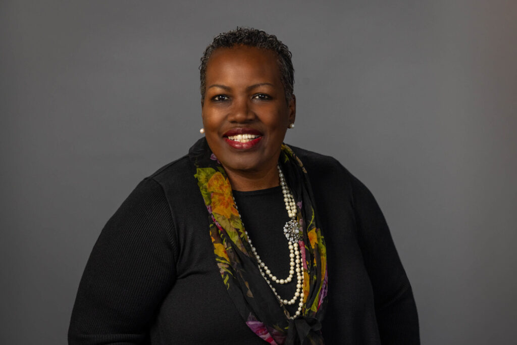 woman in all black outfit with floral scarf and gold jewelry smiles for the camera with confidence