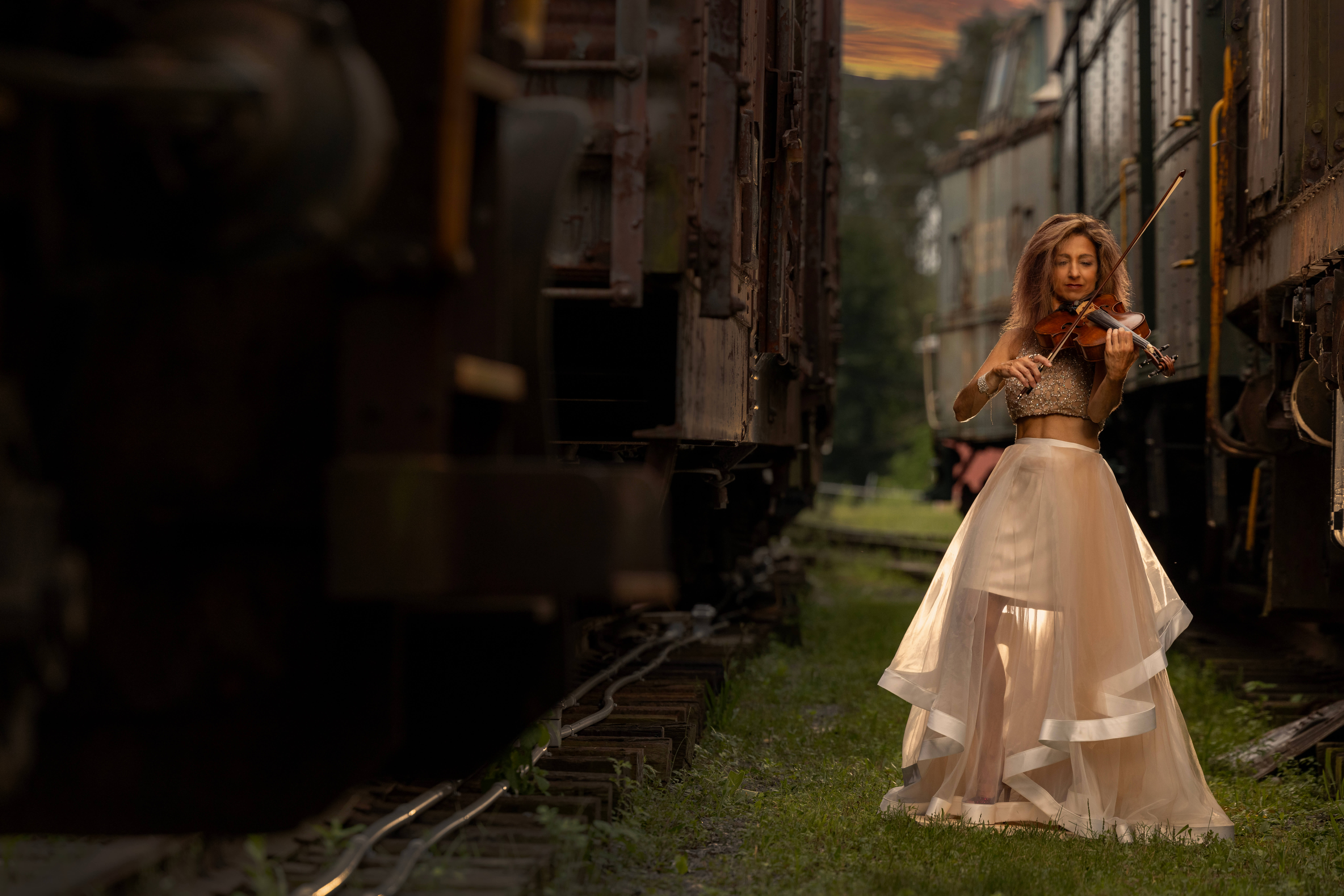 a woman in a flowy dress playing the violin between two abandoned trains