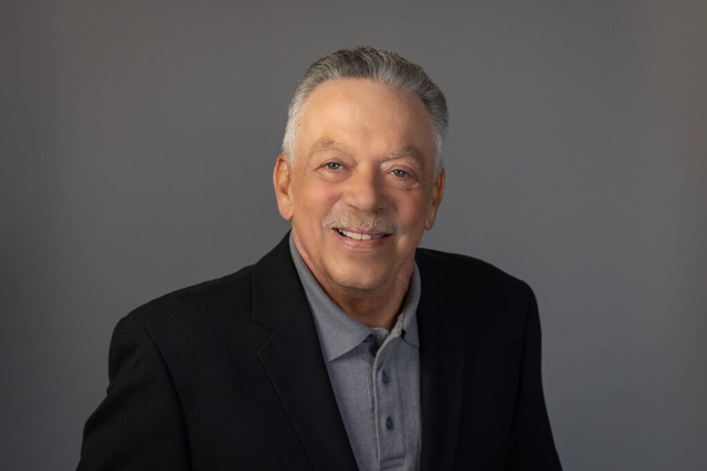 man smiles for camera wearing a button-up shirt and black blazer against grey background