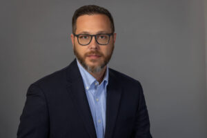 man with serious expression poses in a light blue collard shirt and navy blazer
