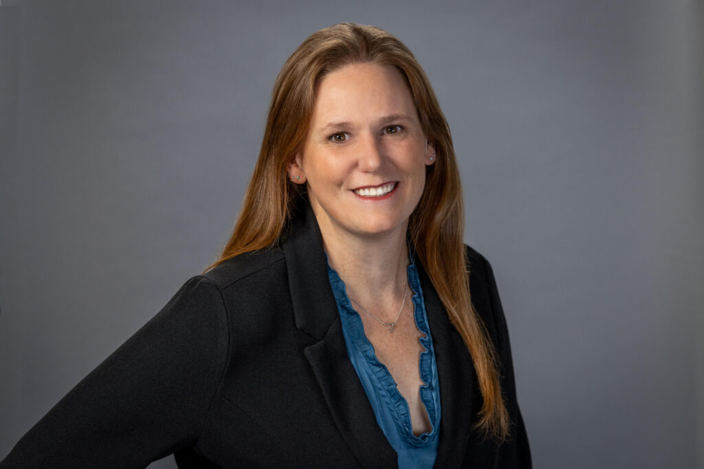 woman poses in teal shirt with black blazer against a grey background in studio