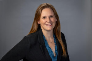 woman poses in teal shirt with black blazer against a grey background in studio