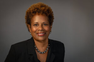 woman with short brown hair against gray background in black suit with jewelry