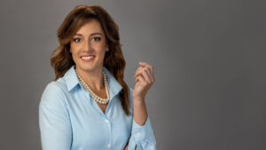 woman with pearl necklace and light blue collared shirt posing with arms half folded