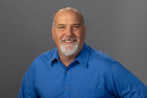 man in blue collared shirt smiles for camera against a grey background