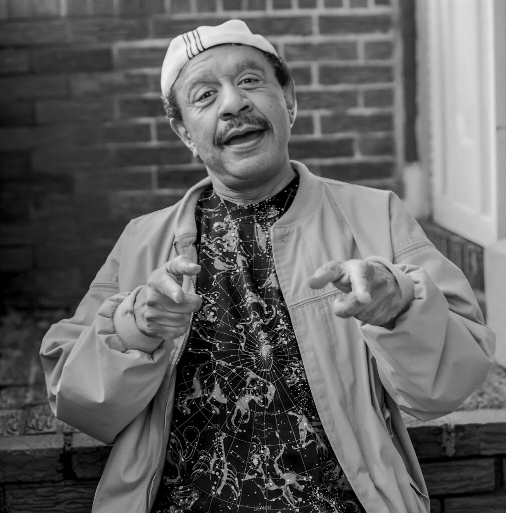 sherman hemsley laughing and pointing at the camera against brick wall