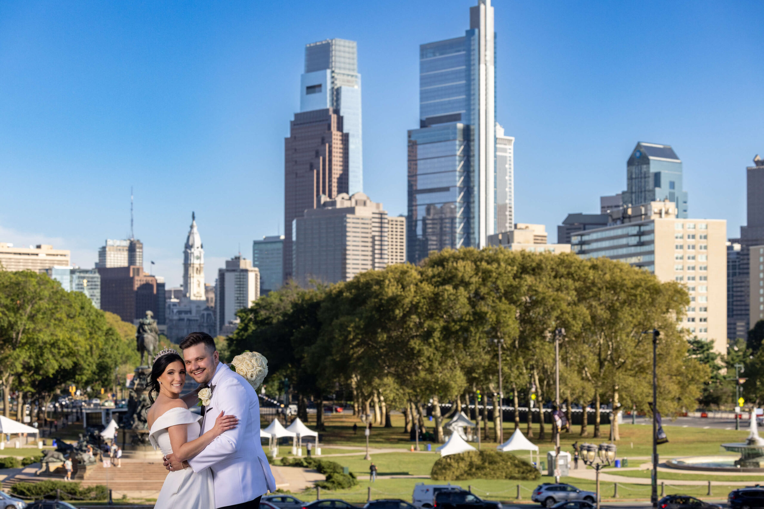 bride and groom posing at art museum with philly skyline behind them on sunny day
