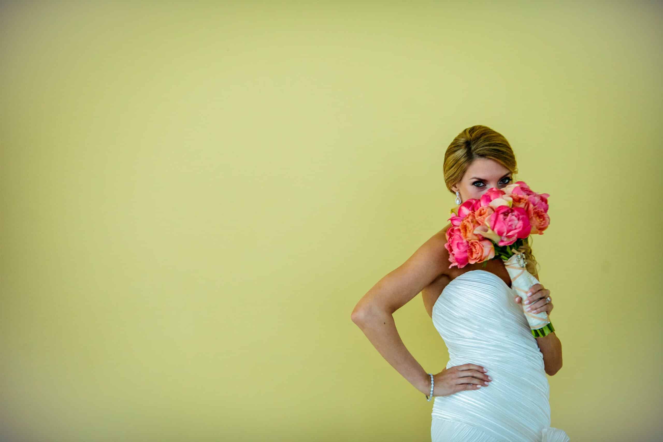 bride hides her face behind a pink flower bouquet against a yellow wall
