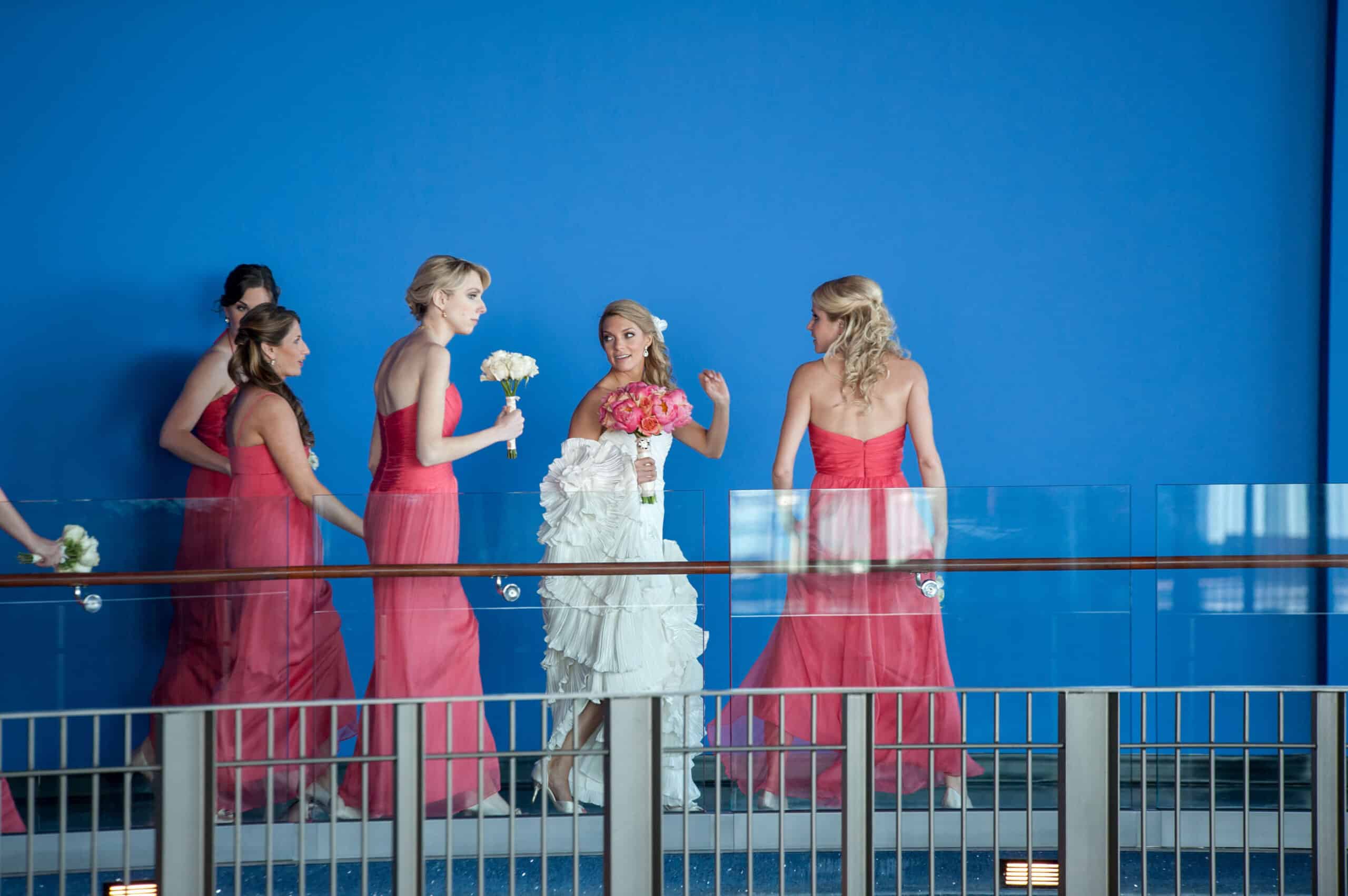 bride and bridesmaids in pink dresses walking against a blue wall