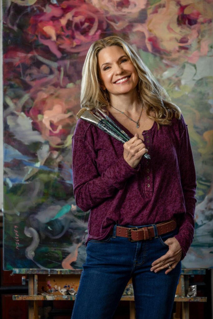 blonde woman in purple shirt holding paint brushes against abstract rose painting