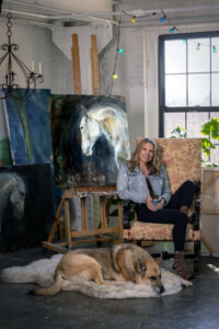 blonde woman in a vintage chair holding a mug in an art studio with a german shepherd dog