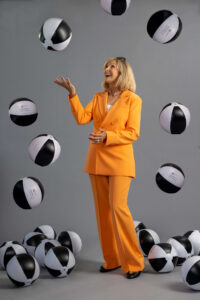 blonde woman in a bright orange suit tossing black and white beach balls into the air and smiling