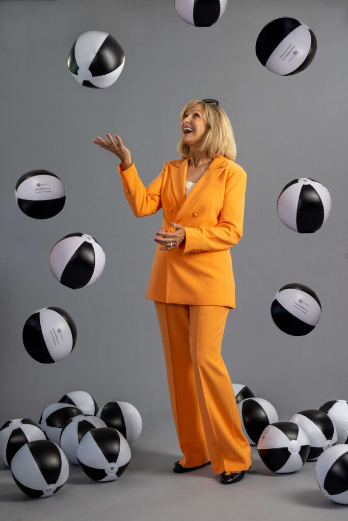 blonde woman in a bright orange suit tossing black and white beach balls into the air and smiling