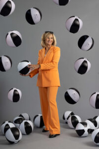 A blonde woman in an orange suit holding and throwing up a bunch of white and black beach balls