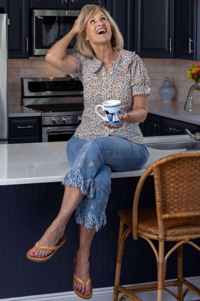 a blonde sitting on a kitchen counter with a cup of coffee in a floral shirt and fringe jeans